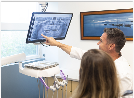 Dentist points to a panoramic dental X-ray on a monitor while explaining it to a patient in a bright, modern clinic; informative tone.