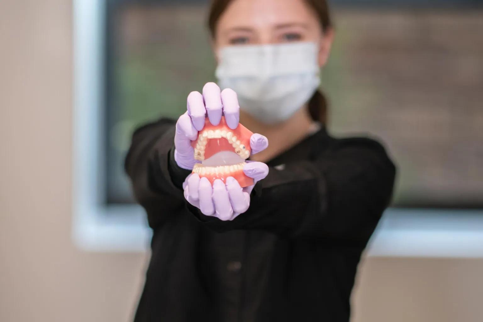 Masked dental professional in black holds a colorful dentures model with gloved hands, illustrating oral healthcare in a clinical setting.