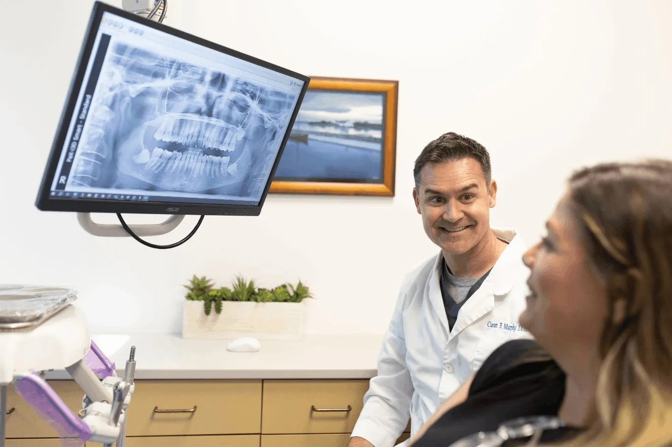 Dentist in a white coat speaks with a female patient in a clinic, beside a panoramic dental X-ray, conveying a calm, attentive consultation.
