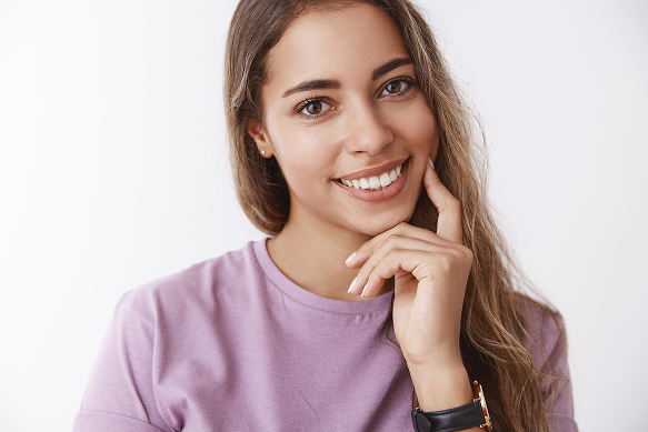 Smiling young woman with long brown hair poses in a lavender shirt, touching her chin against a white background; warm, friendly portrait.